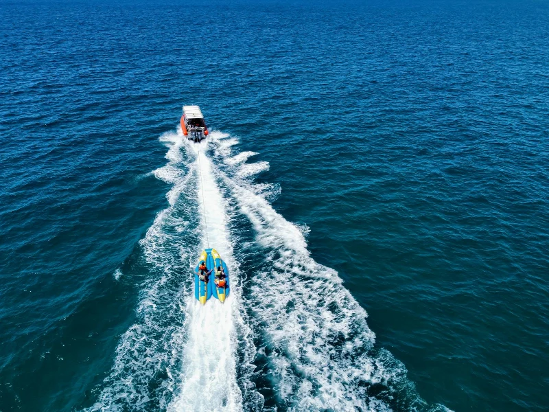 An overhead view of a motorboat towing a banana boat across open blue water